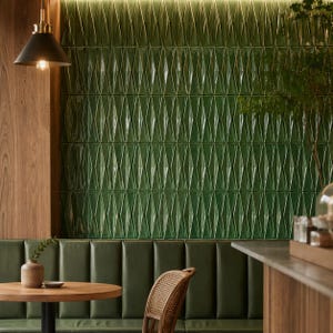 May include: A wall covered in glossy, emerald green, three-dimensional diamond-shaped tiles. The wall is illuminated by a black pendant light fixture. Below the wall is a dark green leather booth, a round wooden table, and a rattan chair.
