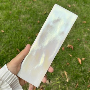 Puede incluir: Una baldosa rectangular iridiscente con un acabado nacarado. La baldosa refleja un espectro de colores, incluyendo blanco, amarillo y verde, creando un efecto brillante. La baldosa se sostiene sobre un fondo de hierba verde.