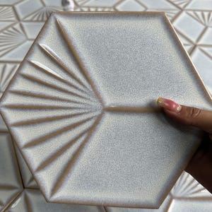 Puede incluir: Una baldosa hexagonal de cerámica con una superficie texturizada y moteada en tonos grises y beige. La baldosa presenta un diseño en forma de abanico que irradia desde una esquina. Los bordes son de un beige más oscuro. Otras baldosas con diseños similares están en el fondo.