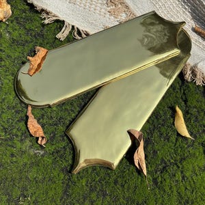 May include: Two gold-coloured, elongated trays with decorative edges are displayed. The trays are positioned on a bed of green moss, with several dried leaves scattered around. A beige woven textile is visible in the background.