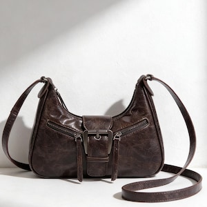 以下が含まれることがあります： カーブした形状と長い調節可能なストラップが付いた茶色のショルダーバッグ。バッグは、シルバーカラーのバックル留めと、前面に2つのジッパーポケットが特徴です。バッグは質感のある素材で作られています。