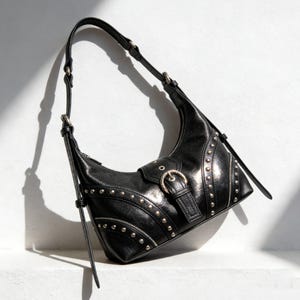 Puede incluir: Bolso de hombro de cuero negro con tachuelas plateadas y cierre de hebilla. El bolso tiene forma curva, una correa larga ajustable y un detalle de cremallera. El bolso está sobre un fondo blanco.