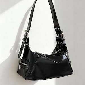 Puede incluir: Bolso de hombro negro brillante con forma rectangular y bolsillo con cremallera en la parte delantera. El bolso tiene una correa larga y ajustable y herrajes plateados. El bolso está hecho de un material brillante.