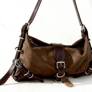 May include: A brown leather shoulder bag with a suede-like texture. The bag features dark brown leather straps, silver metal rings, and buckle closures. The bag has a curved shape and a zipper closure.