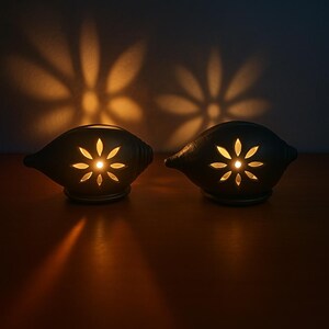 Puede incluir: Dos candelabros de cerámica gris oscuro con forma de concha, cada uno con un recorte en forma de flor que proyecta una sombra floral en la pared. Los candelabros están sobre una superficie de madera, y la luz cálida crea un ambiente acogedor.