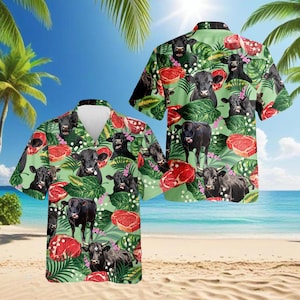 Puede incluir: Camisa hawaiana verde claro con un estampado repetido de vacas negras, filetes rojos y hojas tropicales. La camisa tiene cuello en V y mangas cortas. El fondo muestra una escena de playa con palmeras y cielo azul.