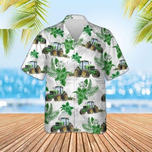 Puede incluir: Camisa blanca de manga corta con un estampado repetido de tractores verdes, follaje verde y flores blancas. Los tractores tienen ruedas amarillas. La camisa tiene un cuello clásico y está hecha de una tela ligera.