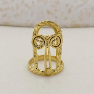 May include: A gold-coloured ring with an ornate design. The ring features a central vertical bar with a decorative swirl at the top and bottom. The band has a textured pattern. The ring is made of metal.