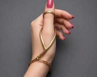 May include: A gold-toned metal hand ornament featuring a ring that encircles the thumb, connected to a curved, Y-shaped structure that extends down to a wrist band. The wrist band includes a short section of flat, interlocking chain links.