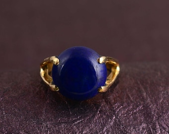 May include: A gold ring featuring a large, oval, deep blue gemstone. The gemstone is the focal point, set in a simple, elegant gold band. The ring is displayed on a textured, dark purple surface.