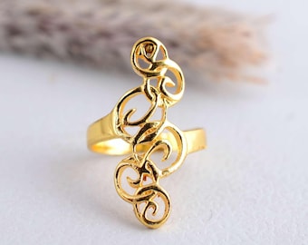 May include: A gold-colored ring with an openwork design featuring swirling, scroll-like shapes. The ring has a band and an elongated decorative element that extends upwards. The ring is set against a soft, blurred background.