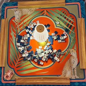 May include: A colorful Mahjong game set with a vibrant orange and teal patterned board. The board features illustrations of cheetahs and tropical leaves. Scattered across the board are numerous blue and white tiles. A decorative lamp is in the center.