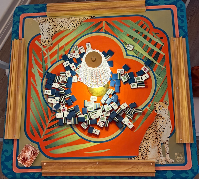 May include: A colorful Mahjong game set with a vibrant orange and teal patterned board. The board features illustrations of cheetahs and tropical leaves. Scattered across the board are numerous blue and white tiles. A decorative lamp is in the center.