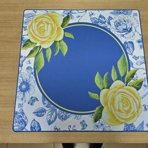 K&ouml;nnte beinhalten: Quadratischer Tischset mit blauem Mittelteil und gelbem Rand. Gelbe Rosen, gr&uuml;ne Bl&auml;tter und blaue Blumenmuster zieren die R&auml;nder. Auf einer hellbraunen Holzoberfl&auml;che platziert.