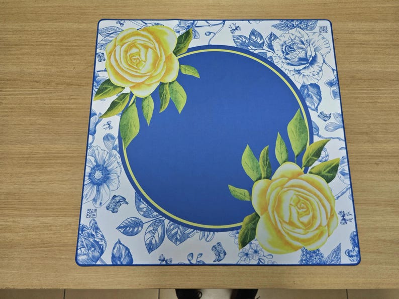 K&ouml;nnte beinhalten: Quadratischer Tischset mit blauem Mittelteil und gelbem Rand. Gelbe Rosen, gr&uuml;ne Bl&auml;tter und blaue Blumenmuster zieren die R&auml;nder. Auf einer hellbraunen Holzoberfl&auml;che platziert.