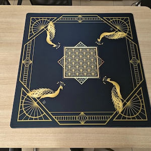 Puede incluir: Una alfombrilla cuadrada de color azul oscuro con un borde geom&eacute;trico dorado y cuatro ilustraciones de pavos reales dorados en las esquinas. Un cuadrado central contiene un patr&oacute;n repetitivo de abanicos dorados y l&iacute;neas geom&eacute;tricas.