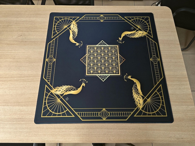 Puede incluir: Una alfombrilla cuadrada de color azul oscuro con un borde geom&eacute;trico dorado y cuatro ilustraciones de pavos reales dorados en las esquinas. Un cuadrado central contiene un patr&oacute;n repetitivo de abanicos dorados y l&iacute;neas geom&eacute;tricas.