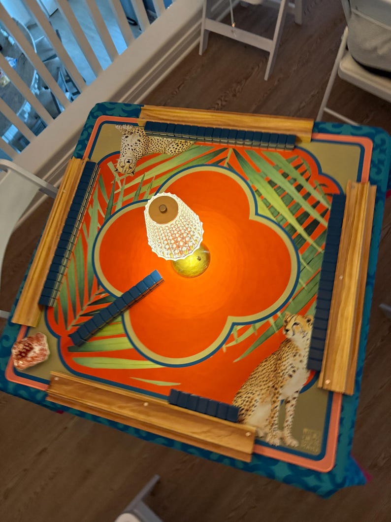 May include: A colorful Mahjong game table with a vibrant orange and teal floral design. The table features cheetah illustrations, wooden rails, and blue tile holders. The game is set up with tiles in place, ready for play.