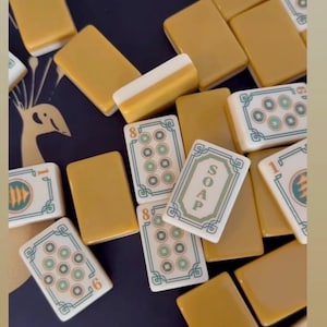 Puede incluir: Una colecci&oacute;n de fichas de Mahjong rectangulares con reverso amarillo mostaza y frente blanco. Algunas fichas presentan patrones circulares verdes y rojos, mientras que otras muestran la palabra SOAP en letras verdes. Las fichas est&aacute;n dispersas.