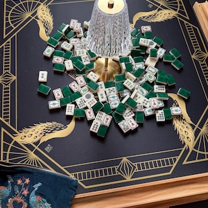 Puede incluir: Una alfombrilla de mahjong negra con ilustraciones de pavos reales dorados, fichas de mahjong blancas y verdes dispersas y una l&aacute;mpara de cristal transparente estriado con base dorada. Incluye un soporte de madera y una bolsa bordada azul.