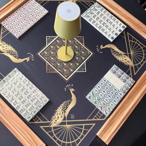 Puede incluir: Una alfombrilla de mahjong negra con ilustraciones de pavos reales dorados y patrones geom&eacute;tricos, que incluye cuatro soportes de madera y juegos de fichas blancas. Una peque&ntilde;a l&aacute;mpara de color verde claro se sit&uacute;a en el centro sobre una mesa redonda.