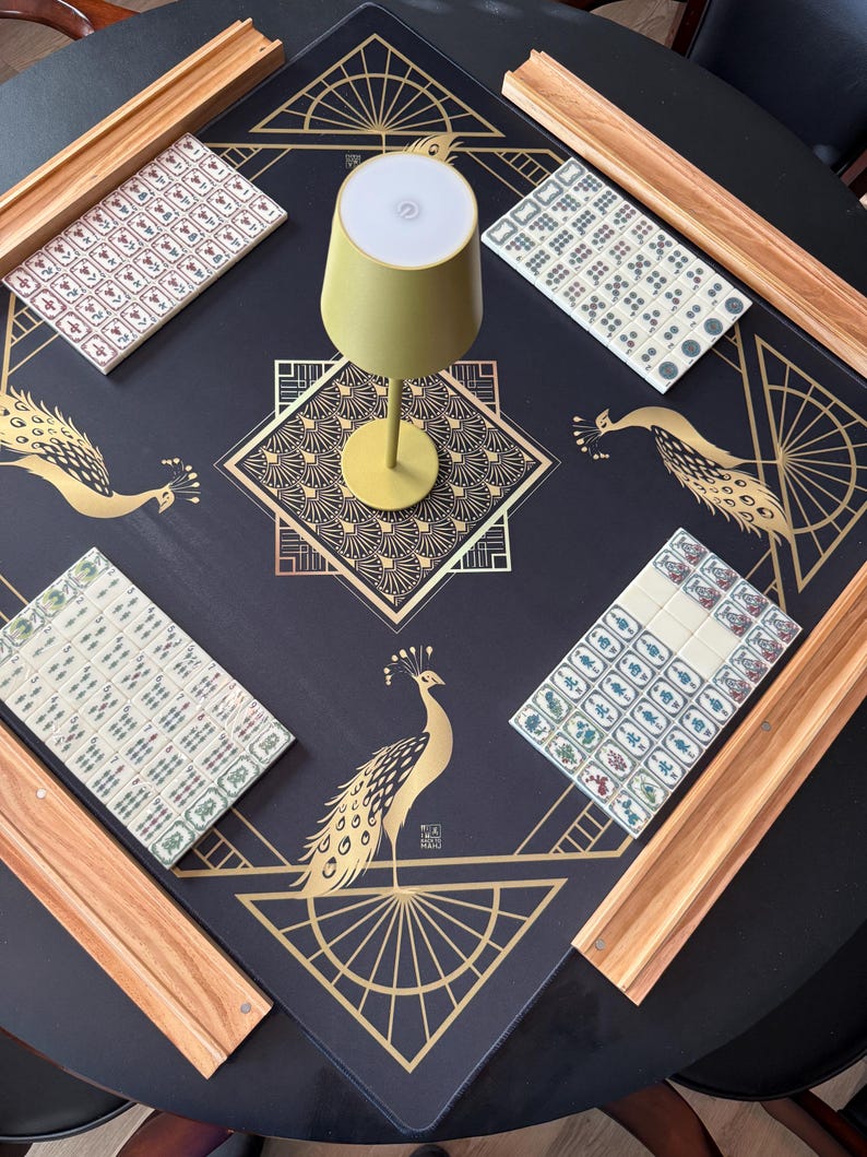 Puede incluir: Una alfombrilla de mahjong negra con ilustraciones de pavos reales dorados y patrones geom&eacute;tricos, que incluye cuatro soportes de madera y juegos de fichas blancas. Una peque&ntilde;a l&aacute;mpara de color verde claro se sit&uacute;a en el centro sobre una mesa redonda.