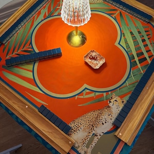 May include: A square Mahjong table with a vibrant orange and teal floral design, featuring a cheetah illustration. The table has wooden edges with blue tile holders. A small lamp and a dish are on the table.