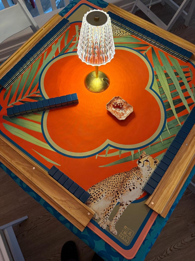 May include: A square Mahjong table with a vibrant orange and teal floral design, featuring a cheetah illustration. The table has wooden edges with blue tile holders. A small lamp and a dish are on the table.