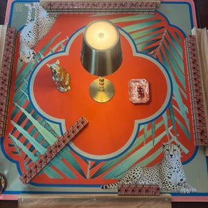 May include: A decorative game board with a vibrant orange and teal design featuring cheetahs and tropical foliage. Includes a gold-colored lamp, a small decorative dish, and a gold-colored cat figurine. The board has wooden rails and game pieces.