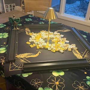 Puede incluir: Un juego de mahjong con fichas amarillas esparcidas sobre un tapete negro con ilustraciones de pavos reales dorados y cuatro soportes negros. Una peque&ntilde;a l&aacute;mpara dorada se encuentra en el centro. El tapete est&aacute; sobre una mesa con mantel de tr&eacute;boles.