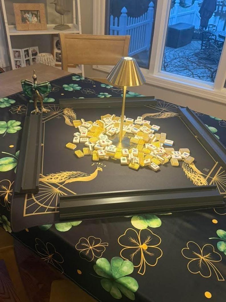 Puede incluir: Un juego de mahjong con fichas amarillas esparcidas sobre un tapete negro con ilustraciones de pavos reales dorados y cuatro soportes negros. Una peque&ntilde;a l&aacute;mpara dorada se encuentra en el centro. El tapete est&aacute; sobre una mesa con mantel de tr&eacute;boles.