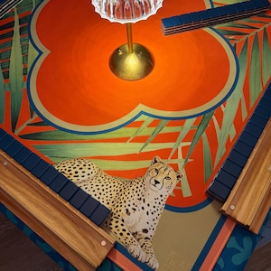 May include: A decorative Mahjong table with a vibrant orange and teal design, featuring a cheetah illustration. The table includes wooden tile holders and a brass lamp. The table's dimensions are approximately 36 inches by 36 inches.
