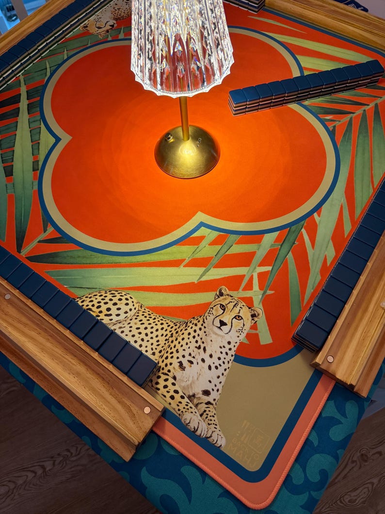 May include: A decorative Mahjong table with a vibrant orange and teal design, featuring a cheetah illustration. The table includes wooden tile holders and a brass lamp. The table's dimensions are approximately 36 inches by 36 inches.