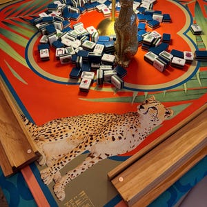 May include: A Mahjong game set on a table with a red and green patterned surface. The set includes blue and white tiles, wooden rails, and a gold-colored cat statue. The table has a cheetah illustration and the text "BACK TO MAHJ".