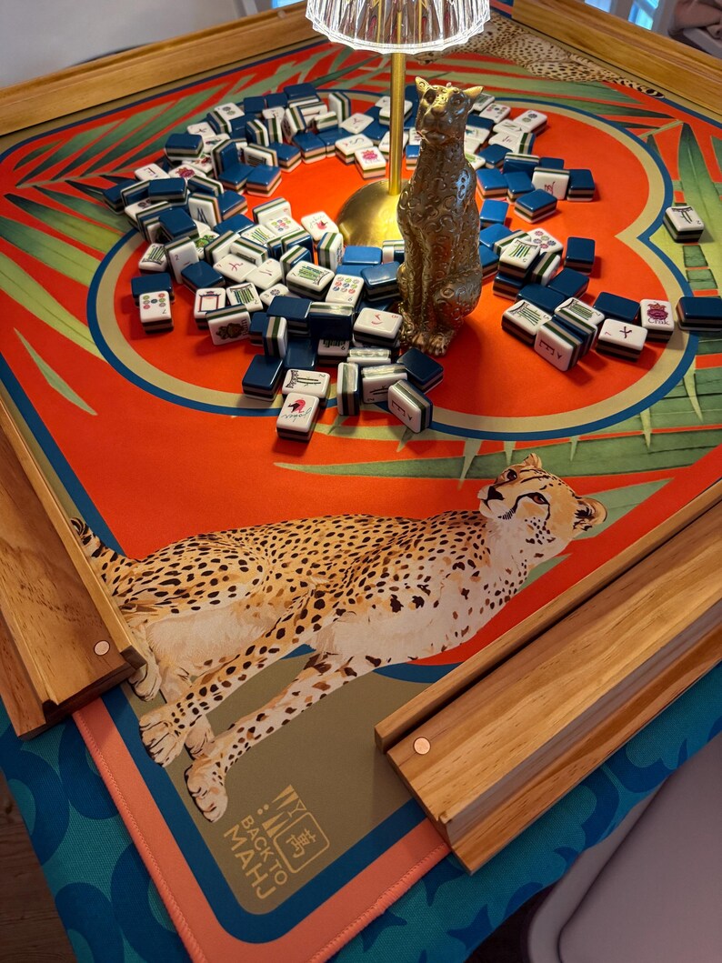 May include: A Mahjong game set on a table with a red and green patterned surface. The set includes blue and white tiles, wooden rails, and a gold-colored cat statue. The table has a cheetah illustration and the text "BACK TO MAHJ".