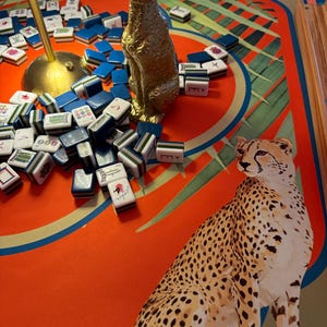 May include: A gold-colored leopard statue sits among blue and white Mahjong tiles on a red and green patterned game board. A cheetah illustration is on the board. A lamp is in the background.