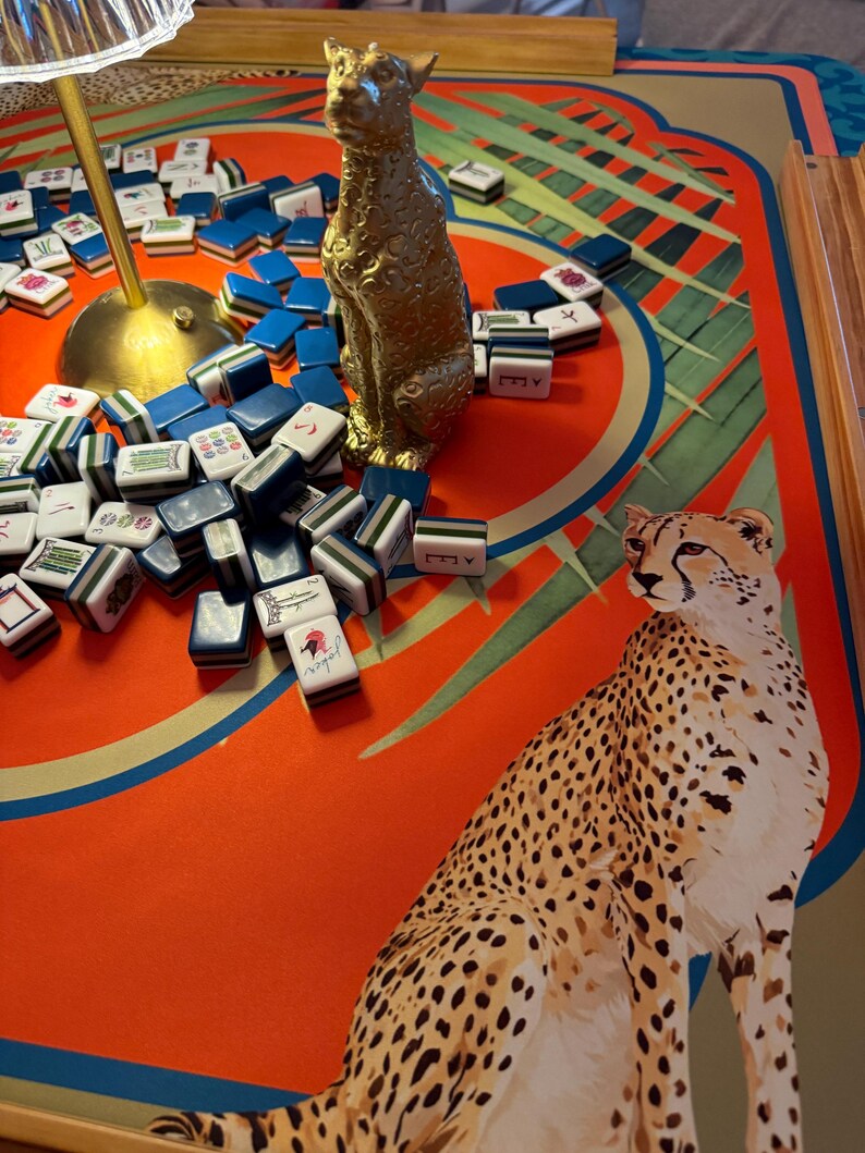 May include: A gold-colored leopard statue sits among blue and white Mahjong tiles on a red and green patterned game board. A cheetah illustration is on the board. A lamp is in the background.