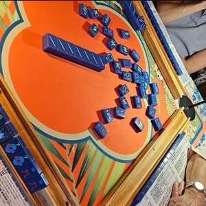 May include: A Mahjong game in progress, featuring blue tiles scattered on an orange and patterned game board. The board has wooden edges and is set up for play. Dice and game pieces are visible, suggesting an active game.