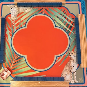 May include: A decorative Mahjong game board with a vibrant design. The board features a central orange quatrefoil shape, surrounded by green palm fronds and cheetah illustrations. Wooden rails and blue tile holders frame the playing surface.