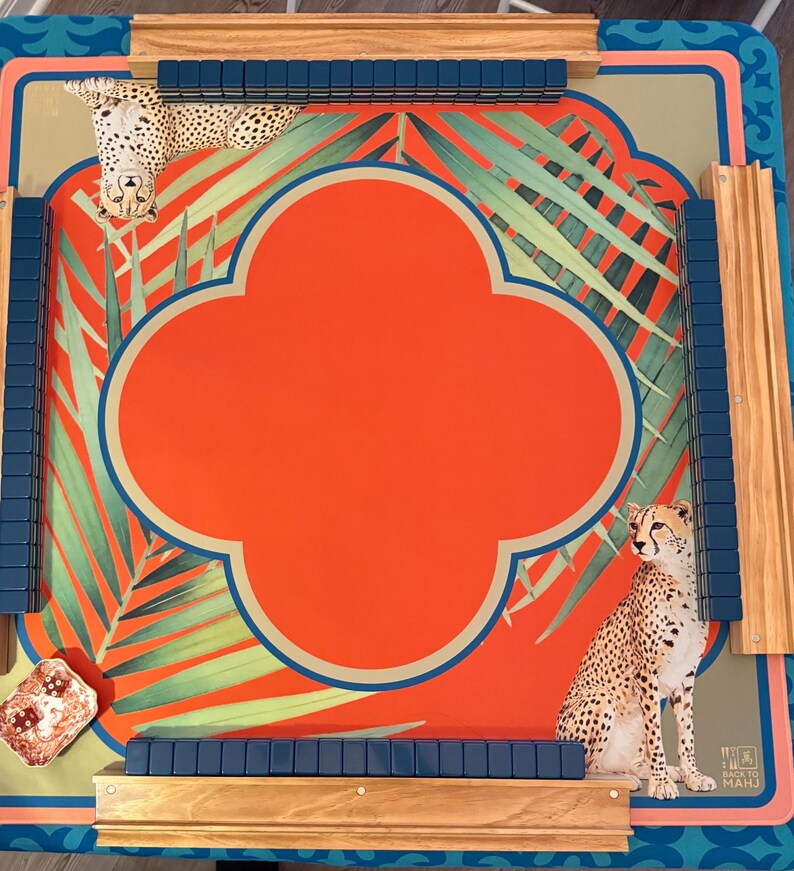 May include: A decorative Mahjong game board with a vibrant design. The board features a central orange quatrefoil shape, surrounded by green palm fronds and cheetah illustrations. Wooden rails and blue tile holders frame the playing surface.