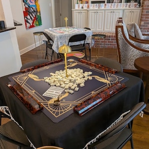 Puede incluir: Una mesa de mahjong cuadrada cubierta con un mantel negro, con una alfombrilla azul oscuro con patrones geom&eacute;tricos dorados y una l&aacute;mpara dorada central. Cuatro soportes de carey rodean un mont&oacute;n de fichas blancas. Un peque&ntilde;o letrero morado dice KEEP CALM AND MAH JONGG.