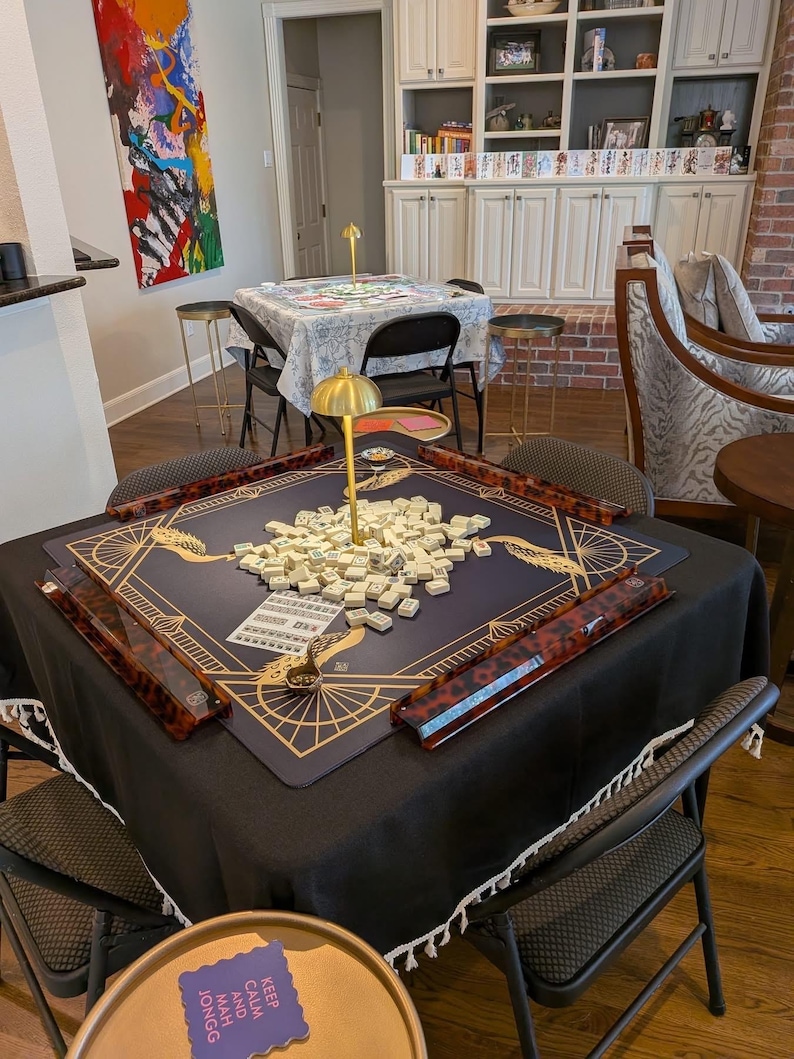 Puede incluir: Una mesa de mahjong cuadrada cubierta con un mantel negro, con una alfombrilla azul oscuro con patrones geom&eacute;tricos dorados y una l&aacute;mpara dorada central. Cuatro soportes de carey rodean un mont&oacute;n de fichas blancas. Un peque&ntilde;o letrero morado dice KEEP CALM AND MAH JONGG.