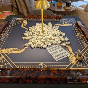 Puede incluir: Una mesa de mahjong cuadrada con superficie de fieltro azul oscuro, ilustraciones de pavos reales dorados y patrones geom&eacute;tricos. Incluye cuatro soportes con estampado de carey, una l&aacute;mpara dorada central, fichas blancas y una hoja de puntuaci&oacute;n.