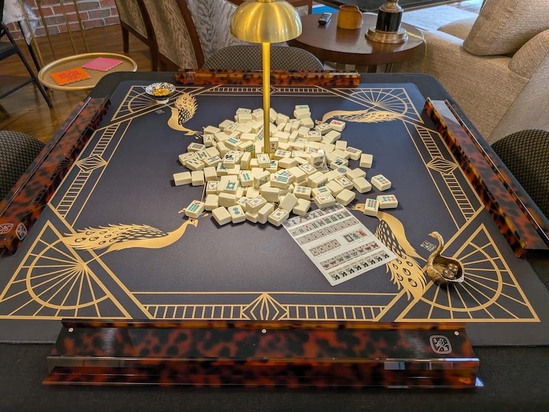 Puede incluir: Una mesa de mahjong cuadrada con superficie de fieltro azul oscuro, ilustraciones de pavos reales dorados y patrones geom&eacute;tricos. Incluye cuatro soportes con estampado de carey, una l&aacute;mpara dorada central, fichas blancas y una hoja de puntuaci&oacute;n.