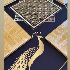 Puede incluir: Una superficie de mesa de mahjong azul oscuro con ilustraciones de pavos reales dorados y patrones geom&eacute;tricos de abanico. Cuatro juegos de fichas de mahjong color crema envueltas en pl&aacute;stico transparente. Se ve el texto grandtilesociety.