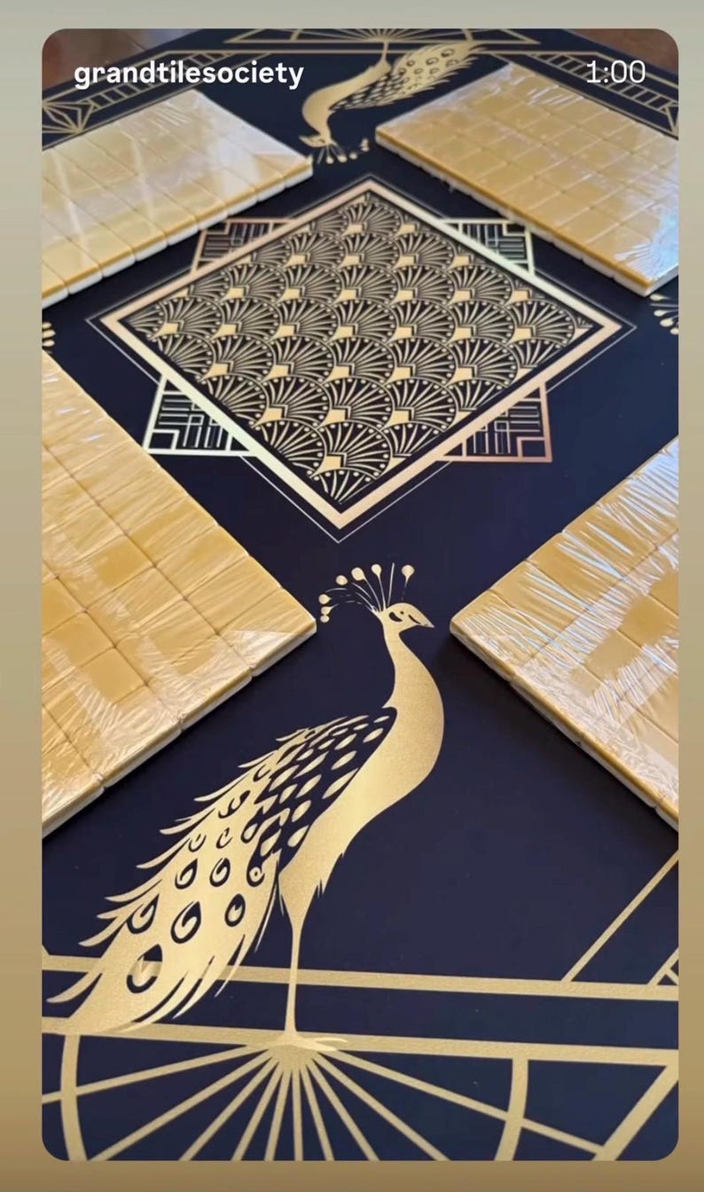 Puede incluir: Una superficie de mesa de mahjong azul oscuro con ilustraciones de pavos reales dorados y patrones geom&eacute;tricos de abanico. Cuatro juegos de fichas de mahjong color crema envueltas en pl&aacute;stico transparente. Se ve el texto grandtilesociety.