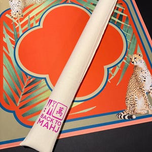 May include: A rolled-up beige Mahjong mat with a matching carrying case. The mat features a vibrant orange cloverleaf design with a teal border and cheetah illustrations. The case has the text "BACK TO MAHJ" in pink.