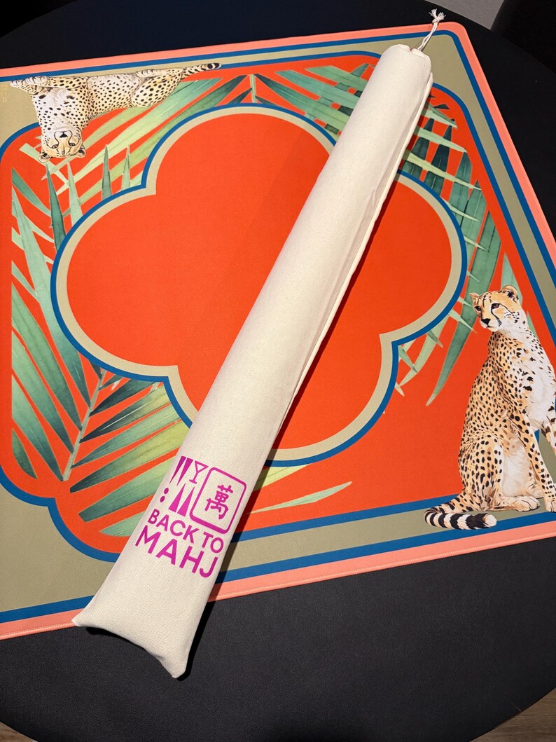 May include: A rolled-up beige Mahjong mat with a matching carrying case. The mat features a vibrant orange cloverleaf design with a teal border and cheetah illustrations. The case has the text "BACK TO MAHJ" in pink.