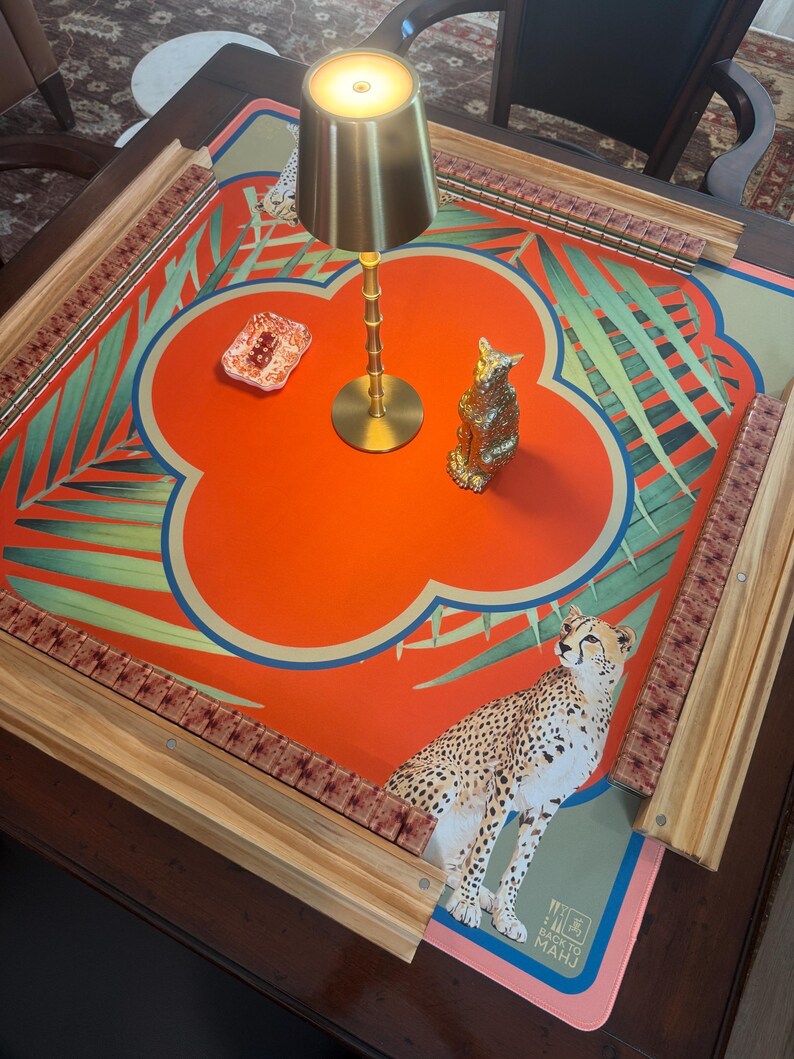 May include: A decorative Mahjong table with a vibrant orange and green floral design, featuring a cheetah illustration. Includes a gold lamp and a small gold cat figurine. The table has wooden edges and a patterned border.
