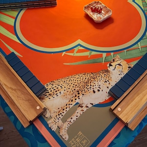 May include: Mahjong set with a vibrant orange and blue floral design, featuring a cheetah illustration. The set includes blue tiles, wooden racks, and a small dish with red dice. The game is set on a patterned mat.